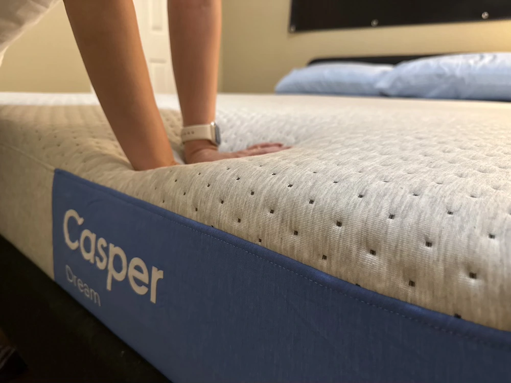 Hands pressing into the Casper Dream mattress, showing compression in its light gray comfort surface