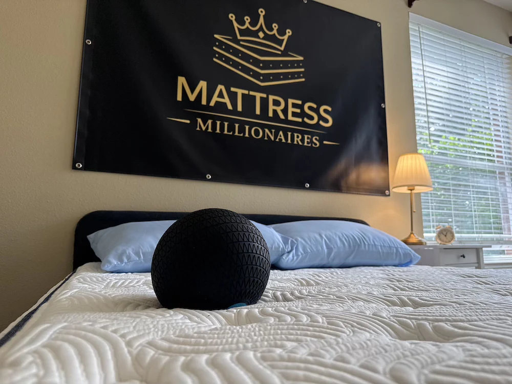 Black textured testing ball resting on a white quilted DLX mattress beneath the Mattress Millionaires sign.