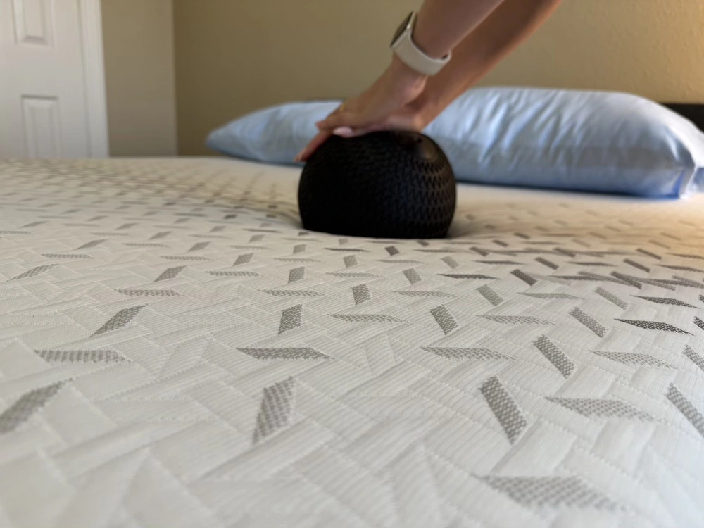 Hands pressing a black textured ball into a white patterned mattress, creating a visible indentation