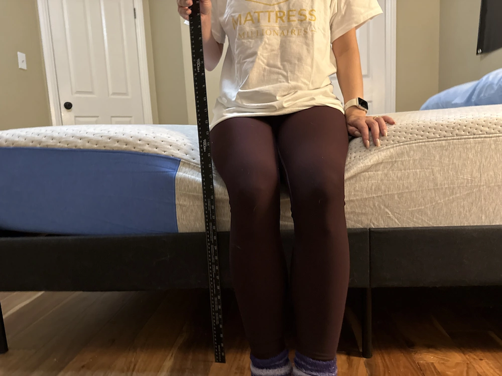 Person measuring mattress sag depth with ruler while sitting on edge of bed