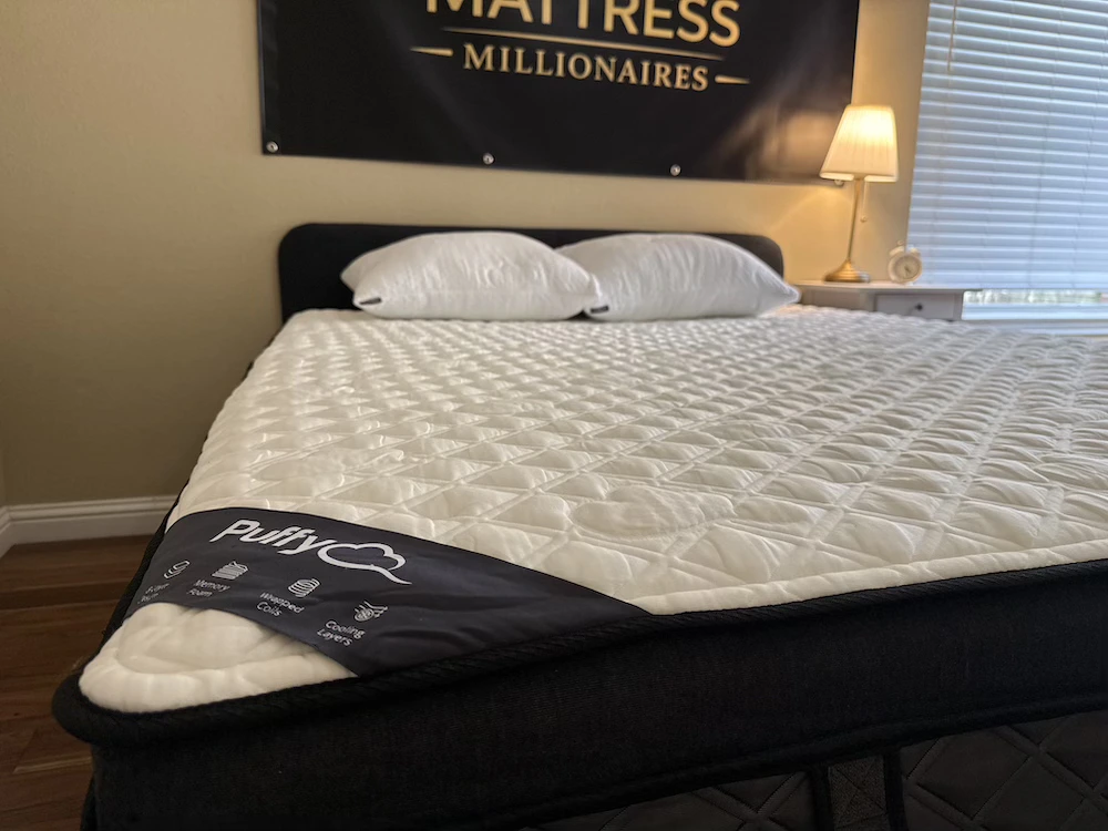 Puffy Lux Hybrid mattress with a white quilted top and dark side panels in the Mattress Millionaires testing room.